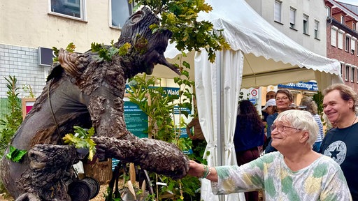 Eine als Baum verkleidete Person gibt einer älteren Frau die Hand