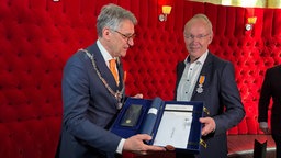 Ein Mann in Anzug, mit Bürgermeister-Kette um den Hals, überreicht einem anderen Mann eine Urkunde