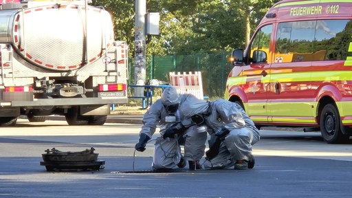 Einsatzkräfte der Feuerwehr knien in Schutzanzügen vor einem großen Gulli und führen mit einem Schlauch Messungen durch