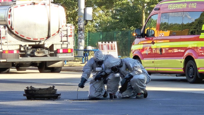 Einsatzkräfte der Feuerwehr knien in Schutzanzügen vor einem großen Gulli und führen mit einem Schlauch Messungen durch