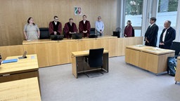 In einem Gerichtssaal stehen acht Personen im Raum. Der Richter liest aus einer Akte vor.