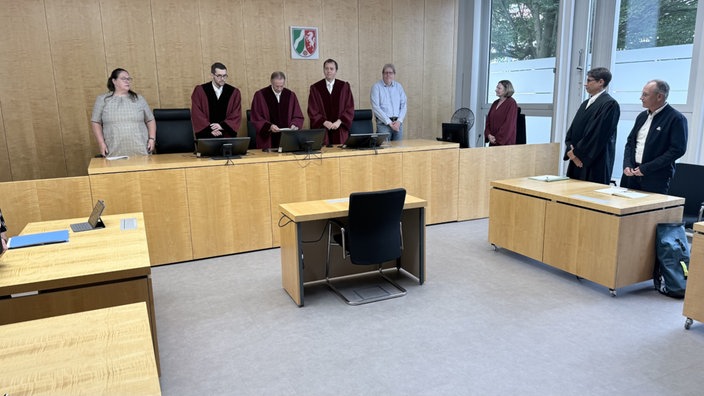 In einem Gerichtssaal stehen acht Personen im Raum. Der Richter liest aus einer Akte vor.