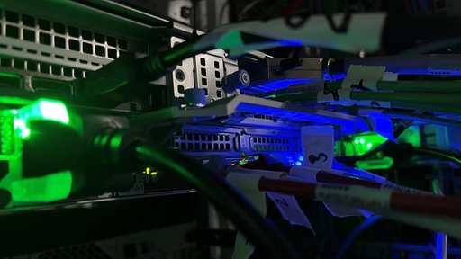 Das Bild zeigt die Nahaufnahme von Kabeln und Eingängen sowie blaue und grüne Lichter an den Apparaturen.