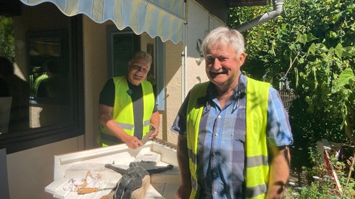 Im Vordergrund steht Paul, dahinter Bert; beide Männer sind in Rente, mit grauen Haaren und einem hellgelben Kittel.