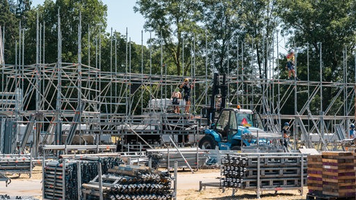 Paletten mit Bauteilen für ein Gerüst, ein Bagger und die ersten Etagen des Baugerüsts sind zu sehen. 