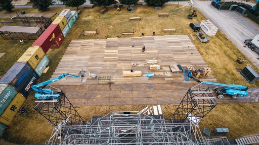 Auf einer Wise wurde eine große Fläche an Holzplatten ausgelegt. Drumherum stehen Container und das Foto wurde von oben von eine Metallbaugerüst geschossen, welches man teilweise sieht. 