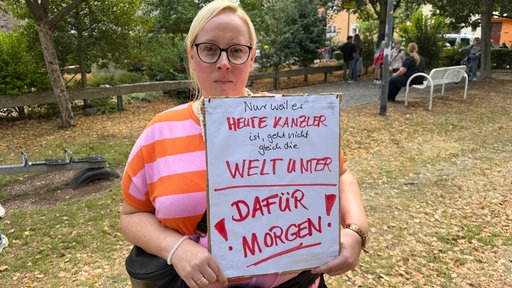 Eine Frau mit buntem Oberteil hält ein Plakat in die Kamera.
