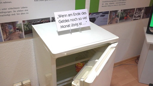 Ein fast leerer Kühlschrank mit offen stehender Türe, darauf ein Schild mit der Aufschrift "Wenn am Ende des Geldes noch so viel Monat übrig ist..."
