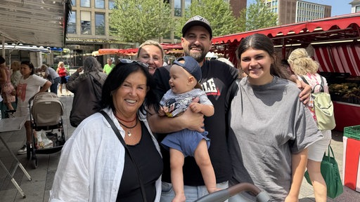 Eine fünfköpfige Familie mit einem Baby steht auf dem Markt und lächelt in die Kamera