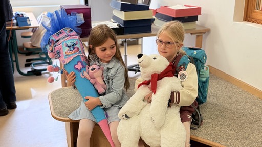 Zwei Mädchen - eine mit Schultüte, eine mit einem Eisbär-Kuscheltier in der Hand - sitzen gemeinsam im Klassenzimmer.