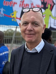 Vor einem Zaun an einem Sportplatz steht ein weißhaariger Mann, dem eine Brille auf der Stirn sitzt.