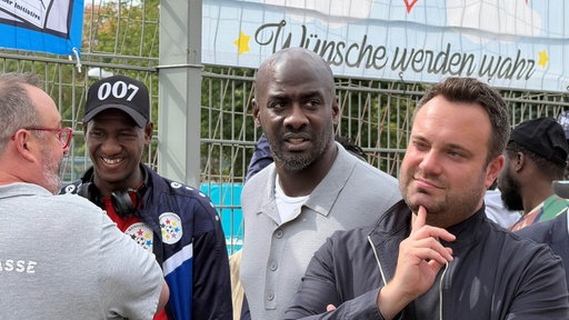 Otto Aldo steht mit anderen Männern in einer Gruppe vor einer Sportanlage. 