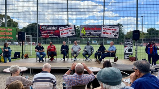 Sieben Personen sitzen auf einer Bühne vor einem Fußballplatz zu einer Podiumsdiskussion zusammen.