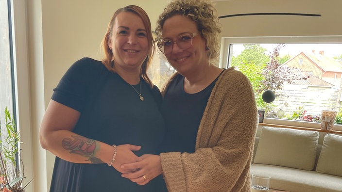 Zwei Frauen stehen dicht nebeneinander. Eine von ihnen ist schwanger. Deshalb hat sie eine Hand auf den Bauch gelegt. Die Frau neben ihr hat wiederum ihre Hand auf die Hand, die auf dem Bauch liegt, gelegt.