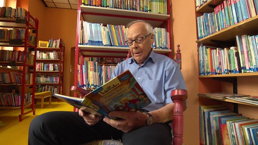 Ein Senior sitzt mit einem aufgeschlagenem Bilderbuch vor einem Bücherregal