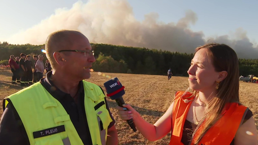 Der Einsatzleiter und die Reporterin auf einem Feld. Im Hintergrund eine Menge Rauch.