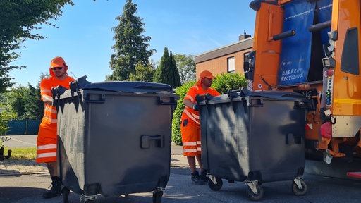 zwei Müllwerker mit Kopfbedeckungen schieben große schwarze Tonnen in Richtung Müllfahrzeug  