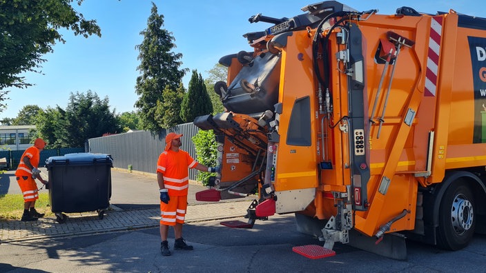 zwei Müllwerker mit orangefarbenen Kappen beladen ein Müllauto mit großen Papiertonnen