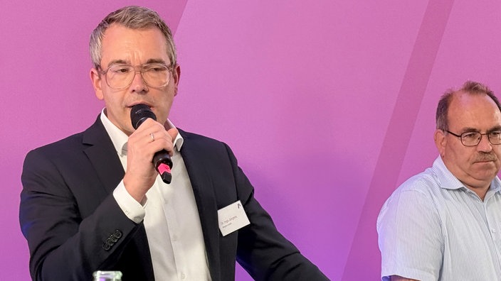 Ein Mann steht vor einem pinken Hintergrund und spricht in ein Mikrofon