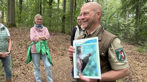 Ranger Sasche Wilden hält ein Bild, auf dem ein Vogel zu sehen ist hoch. Um ihn herum ein paar Personen, die zuhören.