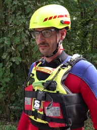 zwei Männer mit neongelben Helmen und Neoprenanzügen stehen an einem Ufer vor Bäumen