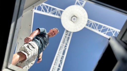 Ein Mann mit Tshirt und kurzer Hose steht unter einem riesigen großen weißen Ding. Vermutlich ist es eine Satellitenschüssel-Konstruktion.