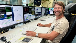 Ein Mann mit blonden Haaren lächelt in die Kamera. Er sitzt an einem Schreibtisch mit ganz vielen Bildschirmen.