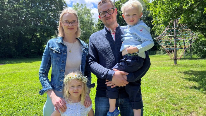 Ein Mann und eine Frau stehen auf einer grünen Wiesen nebeneinander. Der Mann hält einen drei- bis vierjährigen Sohn auf dem Arm. Vor der Frau steht ein ebenso blondes, sechsjähriges Mädchen mit Blumenkranz auf dem Kopf. 