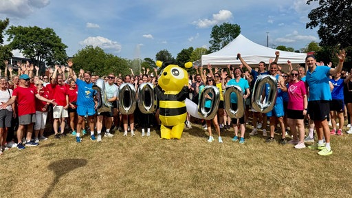 Ganz viele Menschen in Sportkleidung jubeln und halten silberne Luftballons in Form einer 100.000 hoch