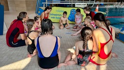 Kinder und Rettungsschwimmer sitzen am Beckenrand und schauen sich Bilder an