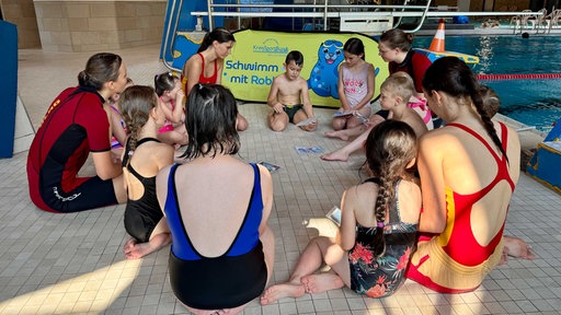 Kinder und Rettungsschwimmer sitzen am Beckenrand und schauen sich Bilder an