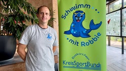 Ein Mann steht neben einem Plakat, auf dem eine Robbe Werbung für Schwimmkurse macht