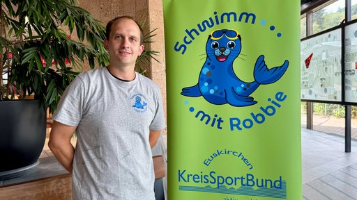 Ein Mann steht neben einem Plakat, auf dem eine Robbe Werbung für Schwimmkurse macht