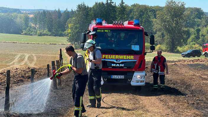 Ein Feuerwehrauto am Feldrand. Davor zwei Einsatzkräfte, die einen Brand löschen.