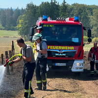 Ein Feuerwehrauto am Feldrand. Davor zwei Einsatzkräfte, die einen Brand löschen.