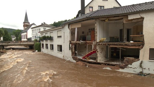 Die Fassade eines Hauses an einem Fluss ist komplett aufgerissen, drinnen sieht man eine Treppe, ein Wohnzimmer und eine Küche