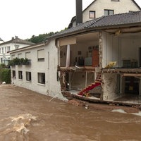 Die Fassade eines Hauses an einem Fluss ist komplett aufgerissen, drinnen sieht man eine Treppe, ein Wohnzimmer und eine Küche