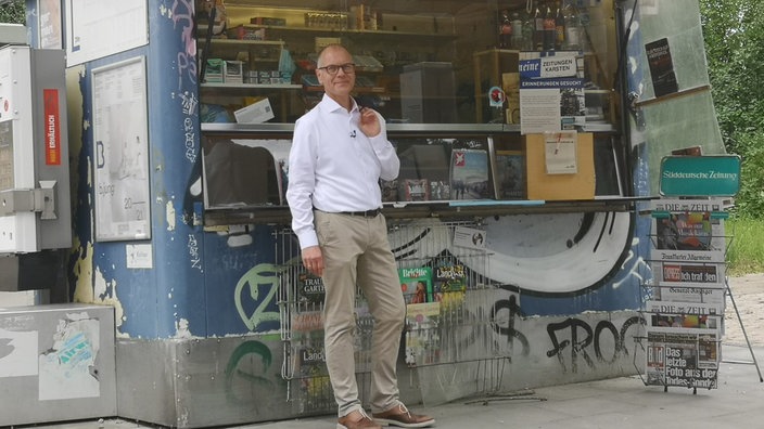 Ein Mann in weißem Hemd steht vor einem alten Zeitungskiosk.