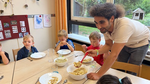 An einem Tisch sitzen mehrere Kinder. Ein Mann tut ihnen aus einer großen Schüssel Nudeln auf ihre Teller.