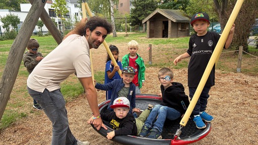 Auf einer Netzschaukel sind vier Kinder. Dahinter stehen noch zwei weitere Kinder. Links an der Schaukel steht ein Mann, der lächelnd in die Kamera schaut.