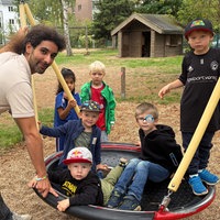 Auf einer Netzschaukel sind vier Kinder. Dahinter stehen noch zwei weitere Kinder. Links an der Schaukel steht ein Mann, der lächelnd in die Kamera schaut.