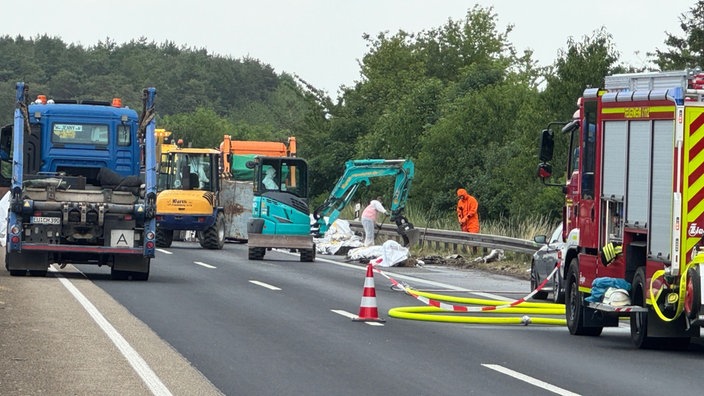 An einem umgekippten Lkw räumt ein kleiner türkiser Bagger mit der Schaufel die verlorene Fracht weg. Zwei Menschen in Schutzanzügen helfen. 