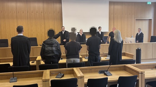 Zwei Männer und eine Frau stehen vor einer Anklagebank