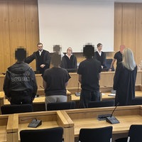 Zwei Männer und eine Frau stehen vor einer Anklagebank