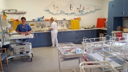 Zwei Schwestern stehen in einem Raum der Geburtsstation. Eine von ihnen versorgt ein Baby, das in einem Bettchen liegt. Im Vordergrund stehen weitere, leere Babybettchen. 