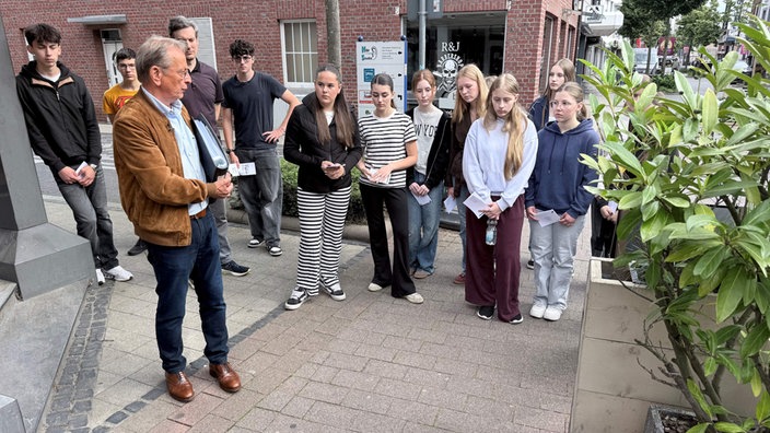 Ein Mann in brauner Jacke steht auf einer Straße einer Gruppe Mädchen und Jungen zugewandt. 