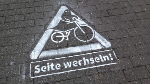 Ein warnendes Dreieck ist mit weißer Farbe auf den Boden gesprüht. In der Mitte vom Dreieck sitzt ein Geist auf einem Fahrrad. Darunter steht "Seite wechseln!".