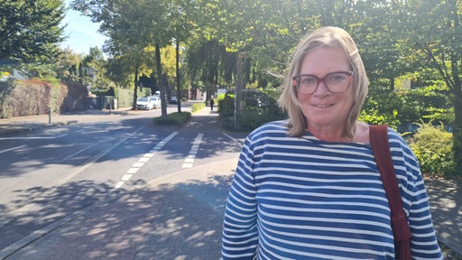 Eine Frau im gestreiftem Oberteil schaut in die Kamera. Im Hintergrund stehen Bäume und links ist eine Straße sichtbar.