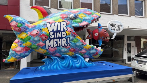 Ein großer Fisch mit der AUfschrift "wir sind mehr" isst einen kleineren Fisch auf dem "wir sind das Volk" steht.