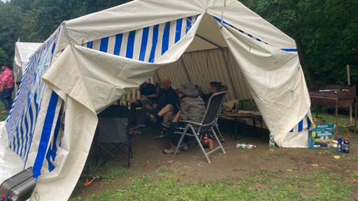 Hinter zwei Flappen eines großen Lagerzeltes sitzen Männer auf Campingstühlen in einem Kreis.